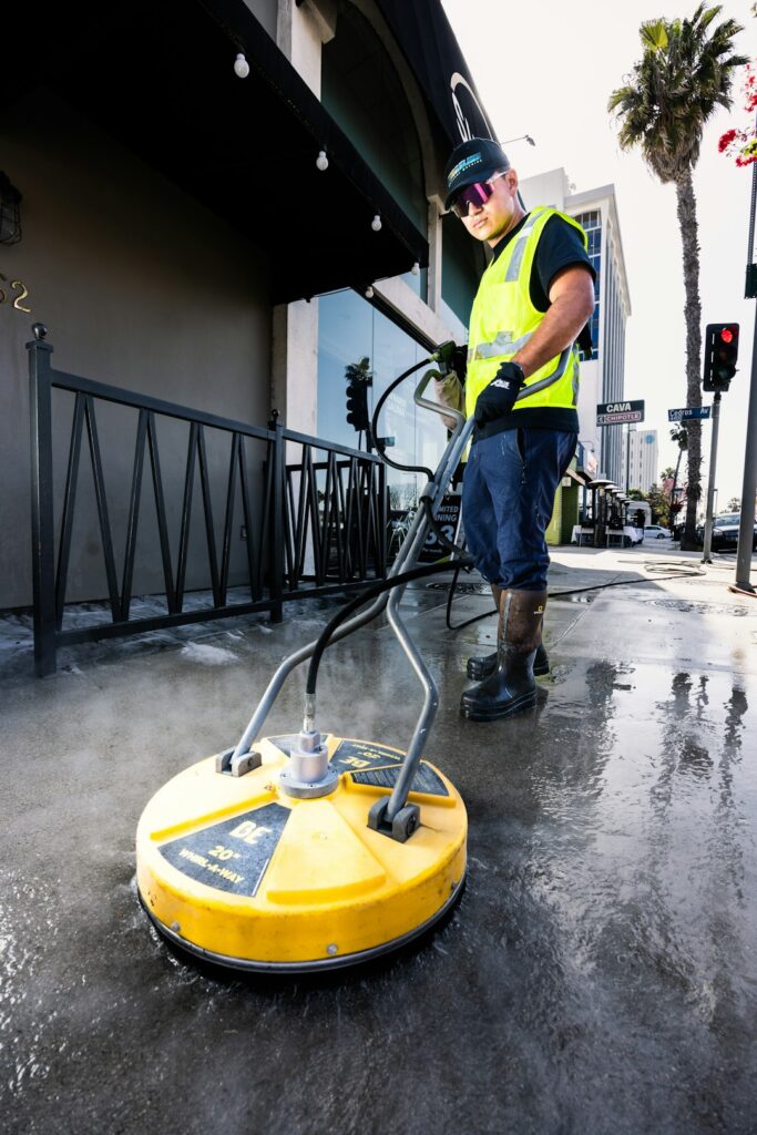 A worker uses a pressure washer on a concrete sidewalk in front of a commercial building.